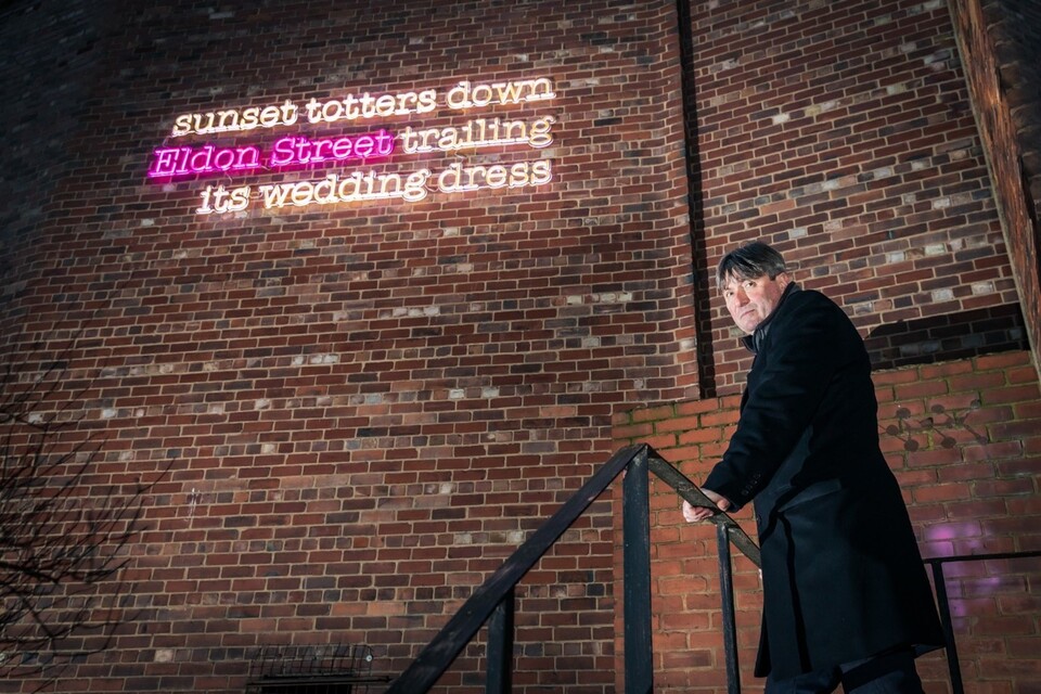 A man in a dark coat stands on outdoor stairs at night, looking at the camera. Behind him, neon text on a brick wall reads: "sunset totters down Eldon Street trailing its wedding dress.